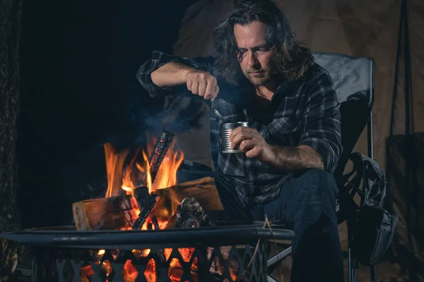 Ateşin yanında kamp yapan bir adam konserve yiyecekler yiyor..