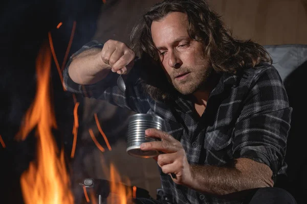 Ateşin yanında kamp yapan bir adam konserve yiyecekler yiyor..