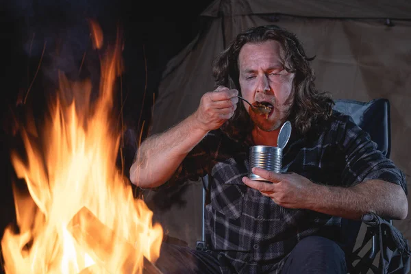 Ateşin yanında kamp yapan bir adam konserve yiyecekler yiyor..