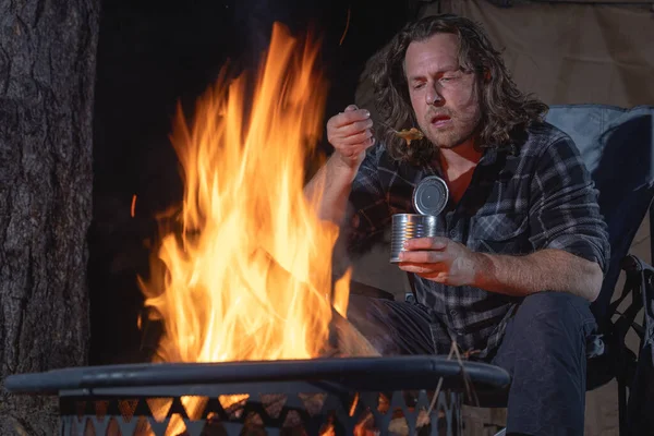 Ateşin yanında kamp yapan bir adam konserve yiyecekler yiyor..