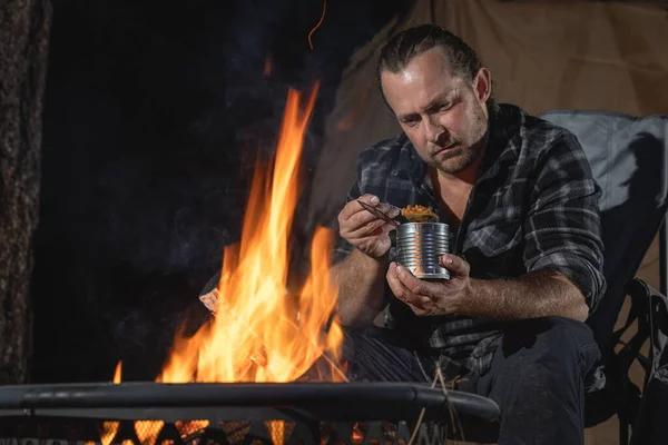 Ateşin yanında kamp yapan bir adam konserve yiyecekler yiyor..