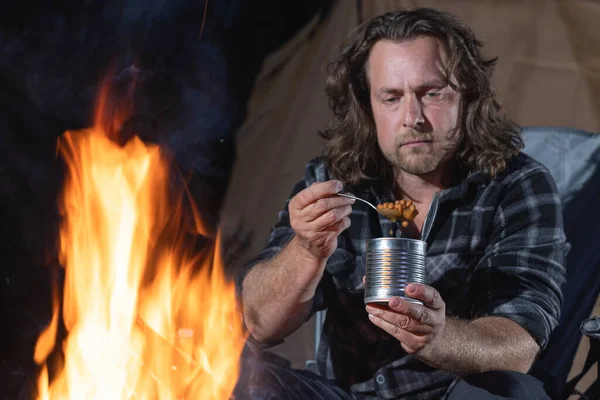 Ateşin yanında kamp yapan bir adam konserve yiyecekler yiyor..