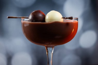 Çikolata karışımının yakın plan bir profili coupe bardakta, gümüş bir kazmada siyah ve beyaz bir çikolata mantarı ile süslenmiş, yumuşak, profesyonel bir bokeh arka planda..