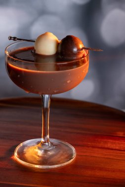 Çikolata karışımının yakın plan bir profili coupe bardakta, gümüş bir kazmada siyah ve beyaz bir çikolata mantarı ile süslenmiş, yumuşak, profesyonel bir bokeh arka planda..