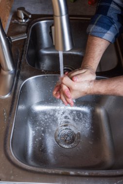 Modern paslanmaz çelik bir mutfak lavabosunda akan suyun altında bir adamın ellerinin ayrıntılı ve eylem odaklı görüntüsü..