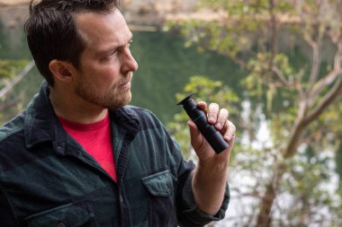 Orta yaşlı ve sakallı bir adamın, arka planda bir göl olan yemyeşil bir ormanda doğayı gözlemlemek için el kameralı siyah bir gözlük kullanmasının yakın plan profili..