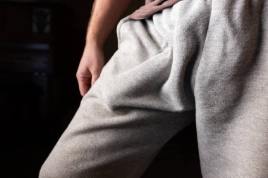 Açık gri kumaş pantolon giyen, göze çarpan bir şekle odaklanan ve cebin içinde koyu bir arka plana doğru şişen bir insanın komik, dar ve detaylı bir görüntüsü..