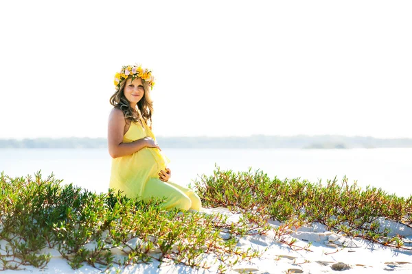 Beautiful smiling pregnant woman with flowers sitting on a beach — Stock Photo, Image