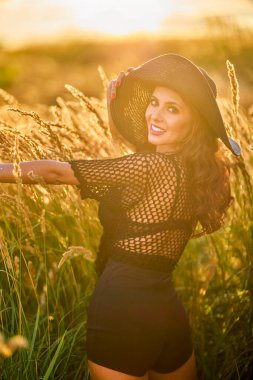 Gün batımında şapkalı güzel bir esmer kadın çimlerde poz verir..