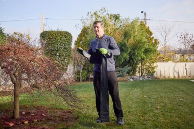 Orta yaşlı bir adam bahçede çalışıyor ve sonbaharda çimleri yapraklardan temizliyor.