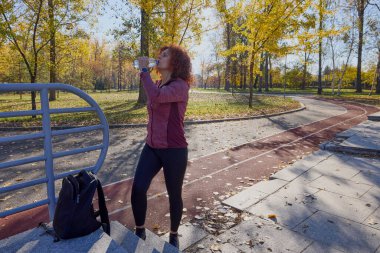 Parkta orta yaşlı bir kadın güneşli bir sonbahar gününde koşu pistinde koşuyor.