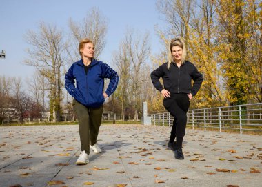 İki orta yaşlı kadın bir sonbahar sabahı parkta ısınma egzersizi yapıyor.