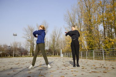 İki orta yaşlı kadın bir sonbahar sabahı parkta ısınma egzersizi yapıyor.