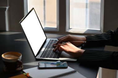 Genç iş kadınının modern çalışma masasında otururken beyaz ekran laptopuyla yazı yazışının yan görüntüsü. Kahve fincanı, grafikli akıllı telefon masanın üzerinde sergileniyor..