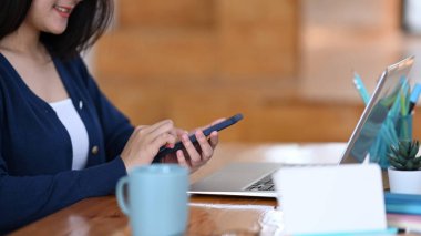 Akıllı telefon tutan ve ofiste otururken dizüstü bilgisayar kullanan bir kadının kesilmiş görüntüsü..