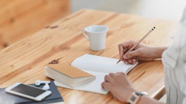 Modern ahşap çalışma masasında otururken çizgili tişörtlü güzel bir kadının yazı / not alışı. Siyah boş akıllı telefon, kahve fincanı ve not defterini masaya koy..