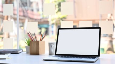 Beyaz beyaz ekranlı bilgisayar dizüstü bilgisayar. Ahşap kalem tutacağı, kahve fincanı, defter ve modern çalışma masasında arka planda ofis camı olan saksı bitkisi..