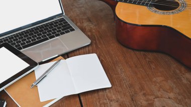 Şarkı sözü yazarının çalışma masası. Kırpılmış nota, kalem, akustik gitar, beyaz beyaz ekran tableti ve dizüstü bilgisayar bunların hepsi ahşap masaya konuyor..