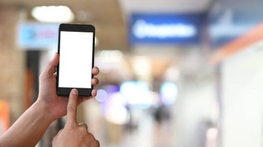 Arkaplan olarak bulanık alışveriş merkezi olan beyaz ekranlı kesilmiş bir siyah akıllı telefonu kullanırken / tutarken yakın çekim elleri.