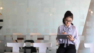 Gözlüklü çekici iş kadını modern ofiste oturuyor ve dijital tablet üzerinde çalışıyor..