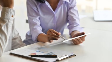 Genç proje yöneticisinin modern çalışma masasında arka plan olarak rahat toplantı odası olan stilus kalem ve tablet kullanarak müşterisine bilgi açıklarken görüntüsü kesildi..