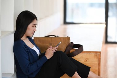 Genç Asyalı kadın, ofiste ahşap zeminde otururken ara verip cep telefonu kullanıyor..