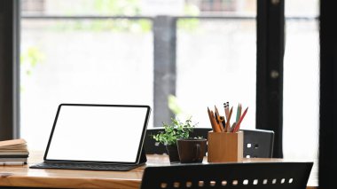 Kalem tutacağı, kahve fincanı ve kitap yığınıyla ahşap masaya yerleştirilmiş beyaz ekranlı bilgisayar tabletinin fotoğrafı. Düzenli ve rahat iş yeri konsepti.