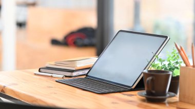 Bilgisayar tabletinin, klavyede beyaz ekranlı fotoğrafı defter, saksı, kahve fincanı ve bulanık arka planı olan modern ahşap masa üzerindeki kalem tutacağıyla bir araya getiriliyor..