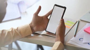 Genç yaratıcı adamın yakın plan görüntüsü çalışan masasında beyaz beyaz ekranlı siyah akıllı telefon tutuyor..