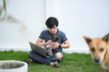 Mutlu genç Asyalı adam arka bahçede çimlerde otururken köpeğiyle oynuyor..