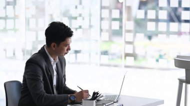 Dizüstü bilgisayarla çalışan ve ofis masasındaki iş verilerini analiz eden kendine güvenen bir iş adamı..