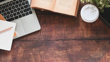Kitap, kalem, kahve fincanı, saksı bitkisi, dizüstü bilgisayar ve defter de dahil olmak üzere ahşap masanın yakın plan görüntüsü..
