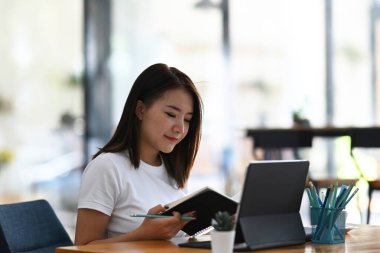 Tablet bilgisayarla çalışan ve ofiste otururken defterden bilgi arayan grafik tasarımcı kadın resmi.