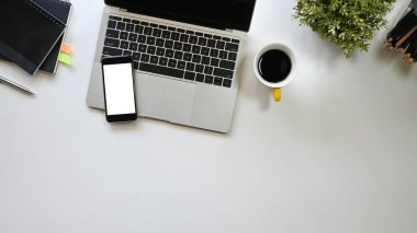 Üst kattaki ofis masasında kahve fincanı, dizüstü bilgisayar, beyaz ekran akıllı telefon, saksı bitkisi, kalem ve defter var. Düz döşeli ofis ekipmanları.