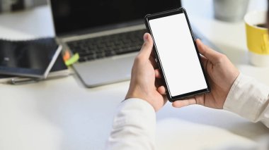 Beyaz ekranlı akıllı telefonu dizüstü bilgisayar, kahve fincanı ve not defterini arka plan olarak beyaz masaya koyarken / tutarken iş adamının kırpılmış görüntüsü.