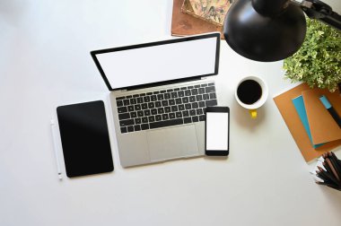 Beyaz beyaz ekranlı dizüstü bilgisayarın üst görüntüsü siyah boş ekran tableti, beyaz ekran akıllı telefonu, not defteri, saksı bitkisi, lamba, kalem tutacağı kahve fincanı ve kitapları bir araya getiriyor..