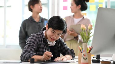 Grafik tablet üzerinde çalışan genç yaratıcı tasarımcı ve arka planda duran meslektaşları.