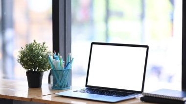 Laptop bilgisayarı, kırtasiye malzemesi, ev bitkisi ve ahşap masadaki defter ile yaratıcı çalışma alanı.