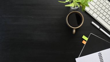 Üzerinde aksesuarlar olan siyah ahşap masa görüntüsü. Düz beyaz klavye, kahve fincanı, saksı çiçeği, defter. Modern ve düzenli iş yeri konsepti.