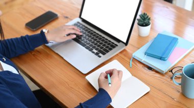 Dizüstü bilgisayarda çalışan ve çalışma alanındaki boş not defterine not alan genç bir kadının yukarıdan çekilmiş fotoğrafı..