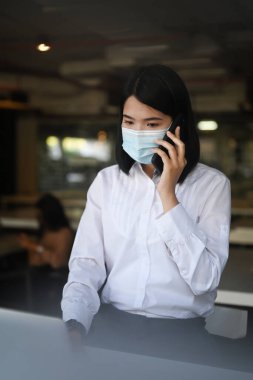 Yüzü maskeli bir iş kadını portresi modern ofiste oturuyor ve cep telefonuyla konuşuyor..