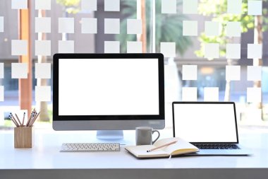 Boş ekran bilgisayarı ve beyaz masada dizüstü bilgisayarı olan modern çalışma istasyonunun ön görüntüsü.