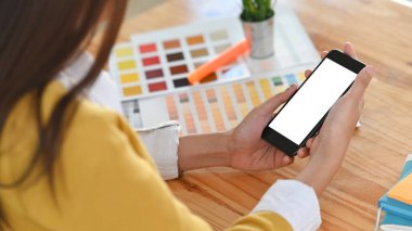 Ahşap çalışma masasının üzerinde elinde beyaz beyaz ekranlı siyah akıllı telefonu tutan genç tasarımcı kızın fotoğrafı..