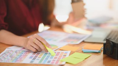 Çalışma masasındaki renk rehberinden renk kodunu seçerken genç grafik tasarımcısının kırpılmış yan görüntüsü. Çalışma masasındaki kamera, kağıt not, dizüstü bilgisayar ve renk rehberi de dahil..