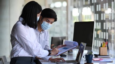 Tıbbi maske takan iki ofis çalışanı modern ofiste proje fikrini tartışıyor..