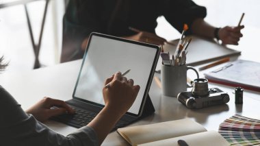 Modern çalışma masasında meslektaşıyla arka plan olarak otururken genç yaratıcı tasarımcının eli beyaz boş ekran tabletine resim çiziyor..