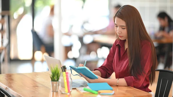 照片上年轻漂亮的设计师穿着红衫 在她的笔记本电脑前 在现代办公背景下的木制办公桌前做笔记 — 图库照片