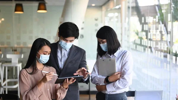 Group of business people in protective mask using digital tablet and discussing business strategy at conference room.