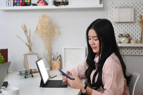 Young Asian woman graphic designer working with tablet computer and using smart phone at her workspace.