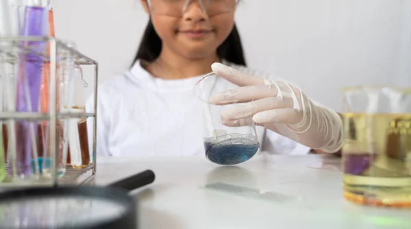 照片中 年轻可爱的女孩戴着安全眼镜和手套 手里拿着一个化学瓶 在一个有着白色背景的现代实验室里做科学实验 — 图库照片